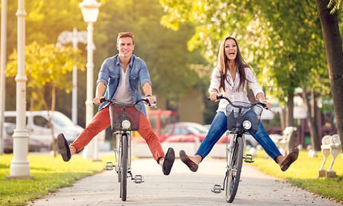 guy and girl being silly with legs outstretched on their bicycles as they ride through a neighborhood park